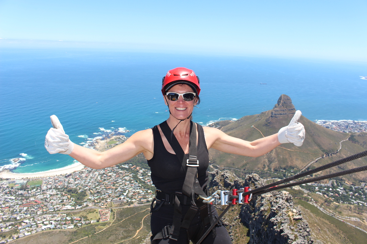 a women with arms outstretched and thumbs up about to abseil down table mountain with a spectacular view of the ocean in the background